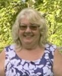 A photo of a woman with grey hair smiling into the camera. She is wearing sunglasses and a purple shirt with white flowers.